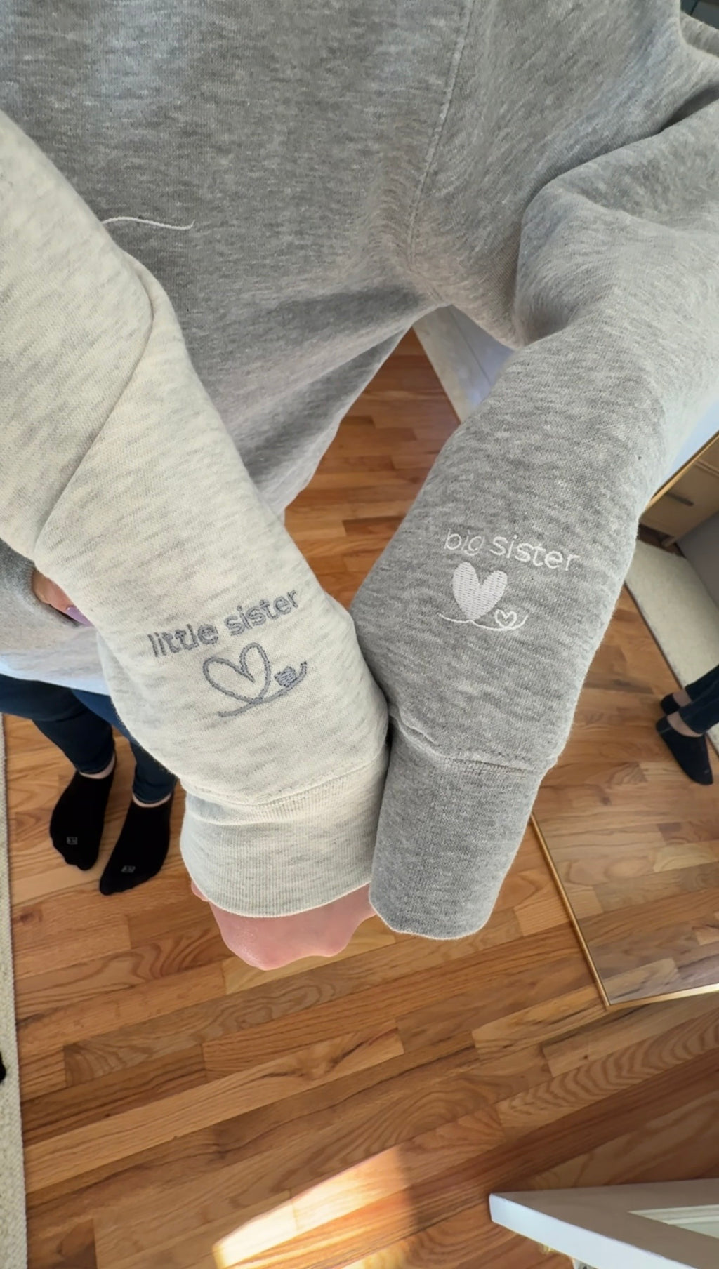 Heather oatmeal sweatshirt with 'little sister' text in grey embroidery on the left sleeve next to a Heather grey sweatshirt with 'big sister' text in white embroidery on the left sleeve