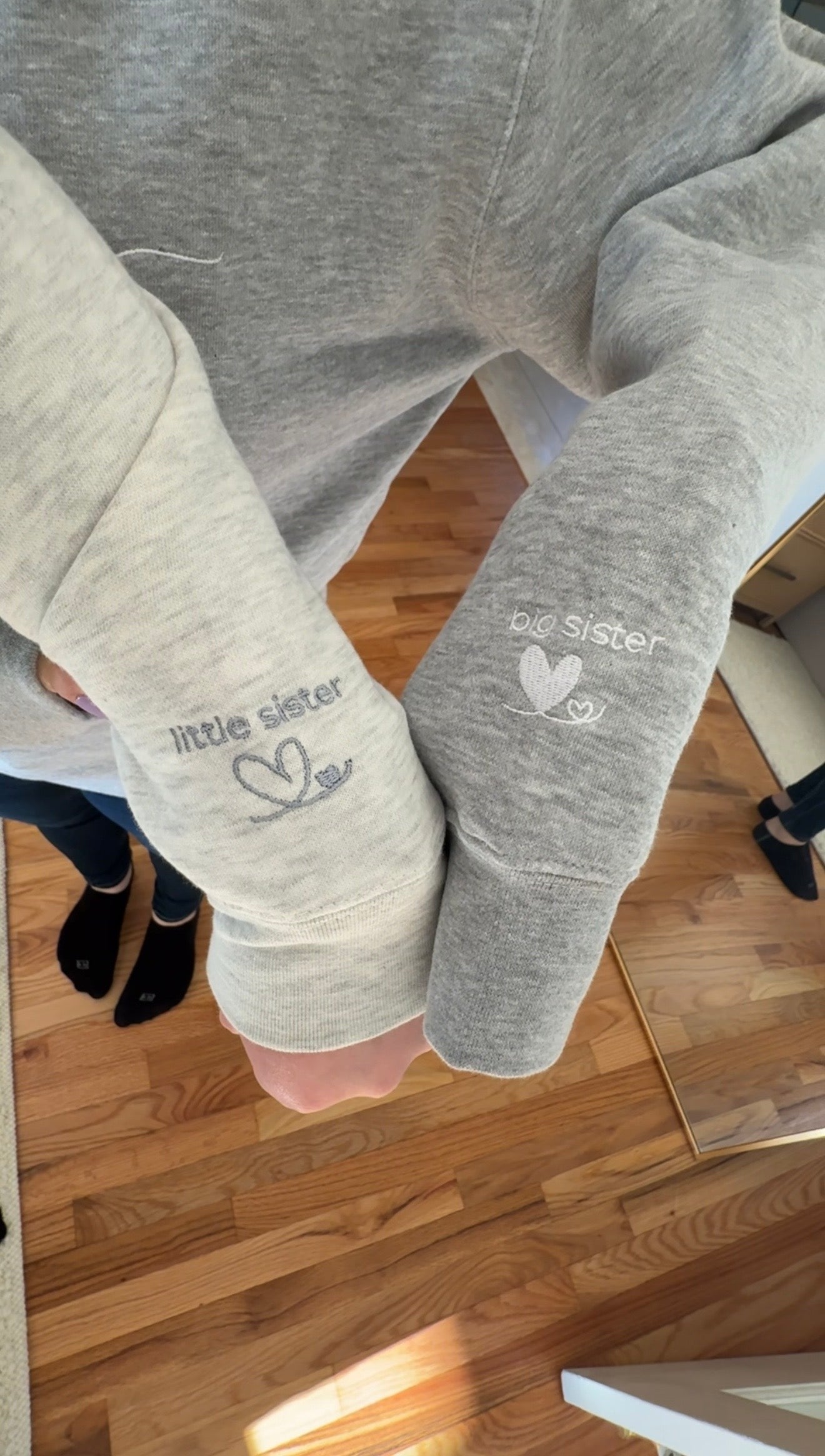 Heather oatmeal sweatshirt with 'little sister' text in grey embroidery on the left sleeve next to a Heather grey sweatshirt with 'big sister' text in white embroidery on the left sleeve