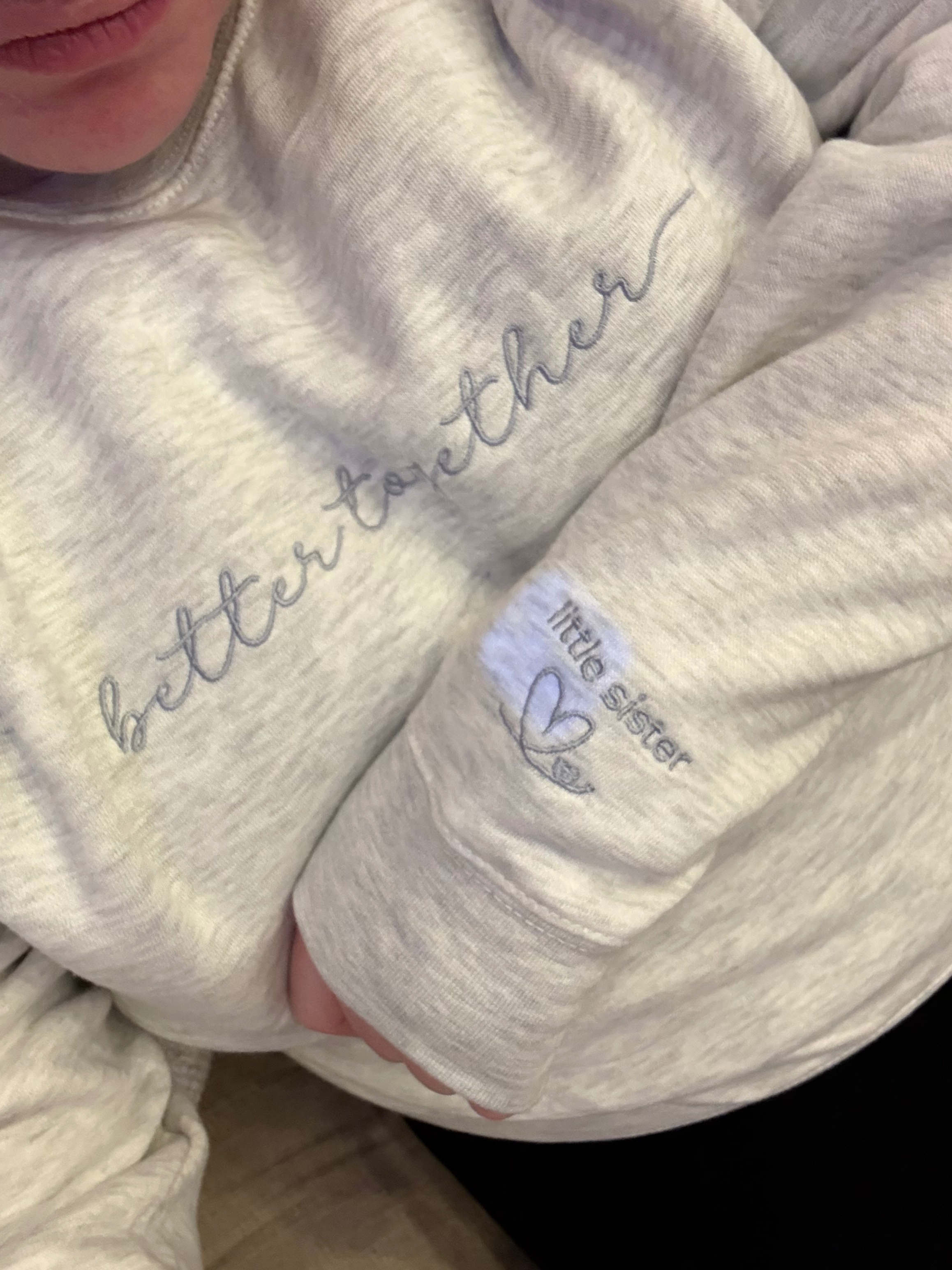 Heather oatmeal sweatshirt with 'better together' text in grey embroidery across center front and little sister with hearts on the left sleeve worn by a person
